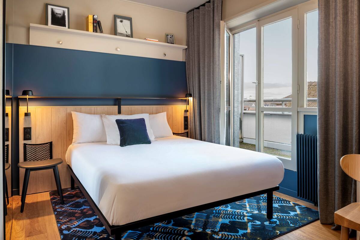 Ein Hotelzimmer mit einem großen Bett, einem Sessel, einem kleinen Tisch, einem Regal mit Gegenständen und einem Fenster mit Vorhängen.