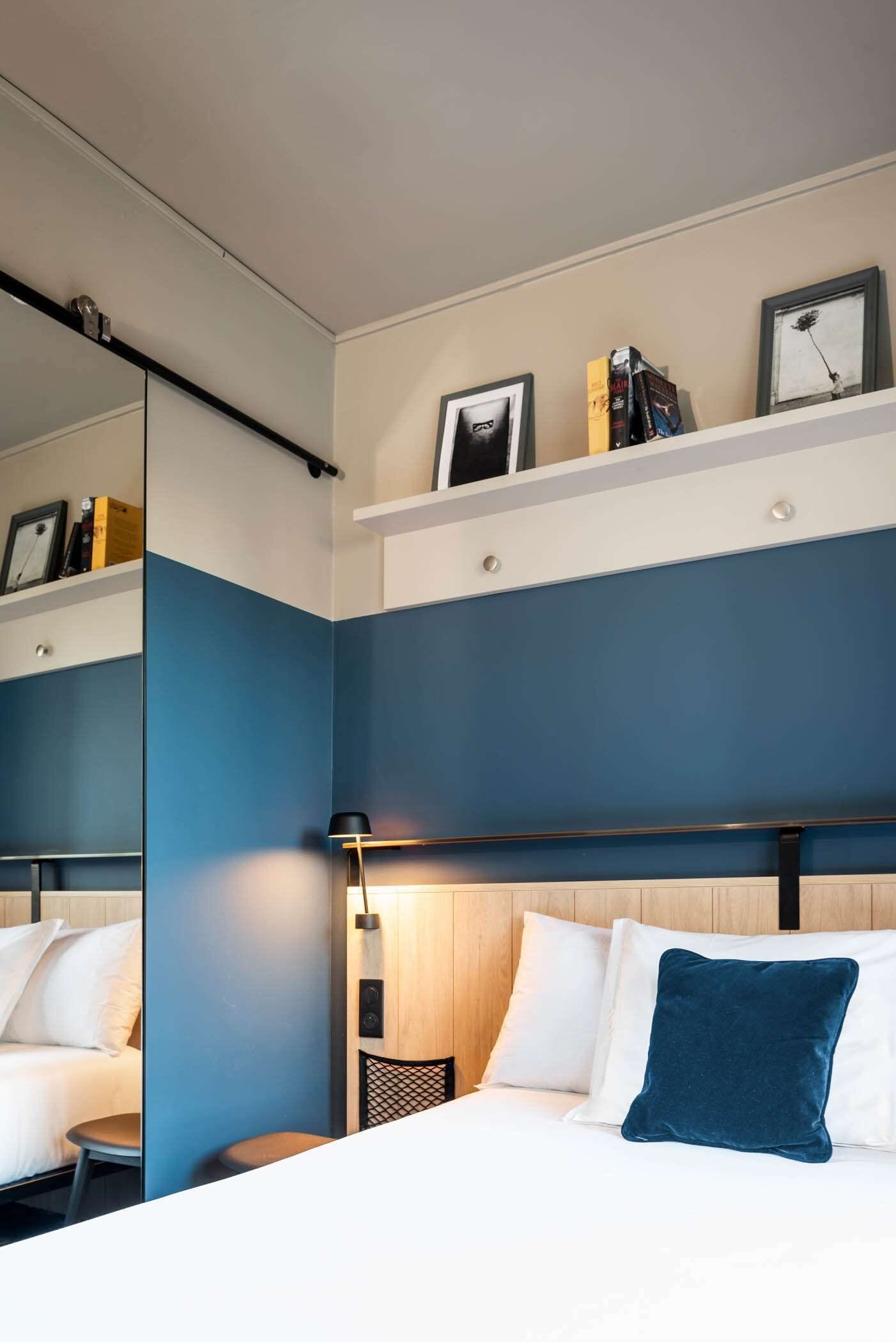 Ein Hotelzimmer mit Bett, Nachttisch, Spiegel und einem Regal mit Büchern und einem Bilderrahmen.
