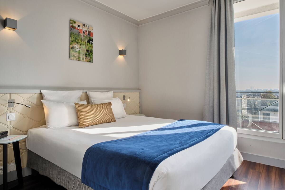 A hotel room with a large bed, a nightstand, a wall-mounted lamp, and a window with a view of buildings.