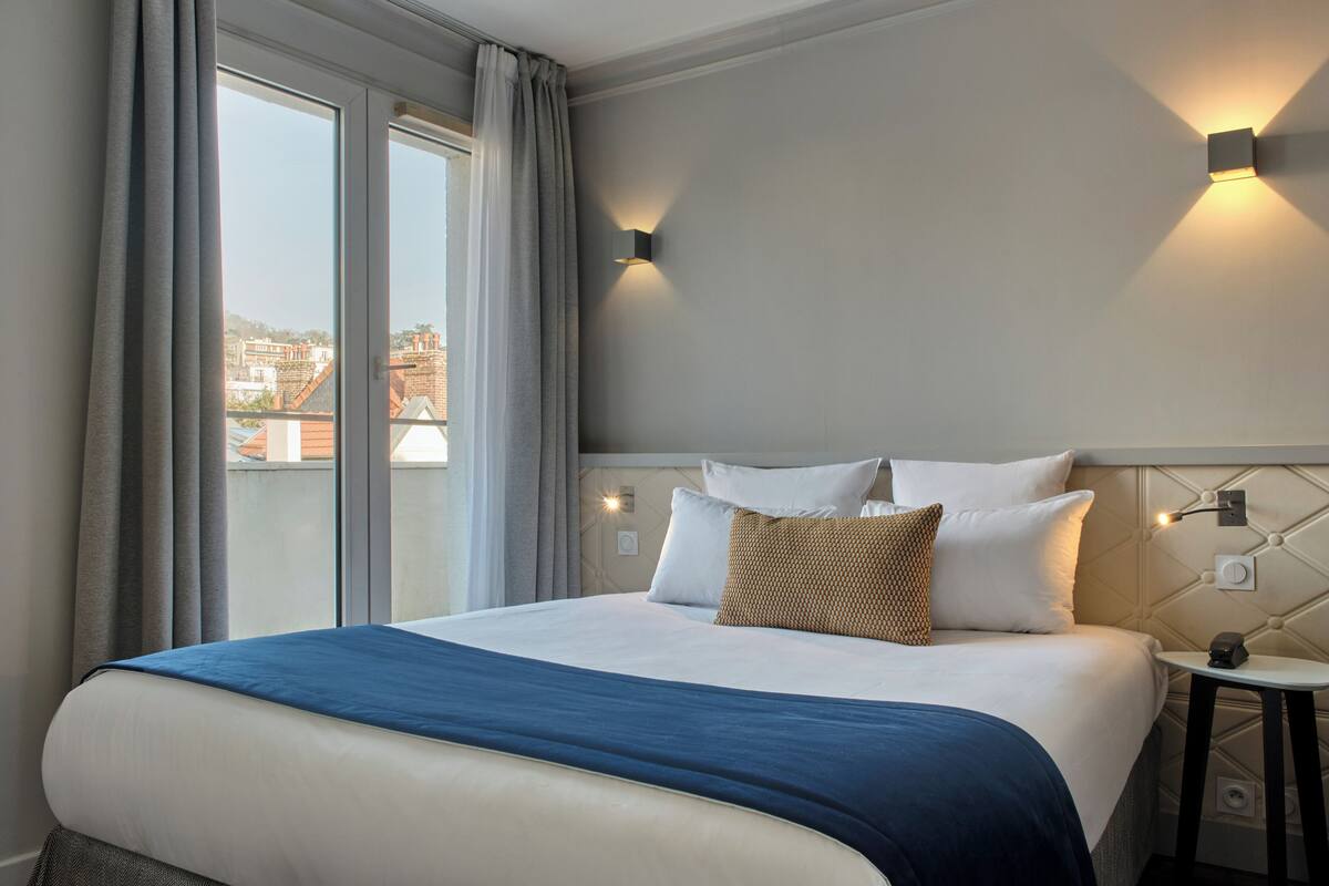 A hotel room with a large bed, a view through a window, and a wall-mounted light fixture.