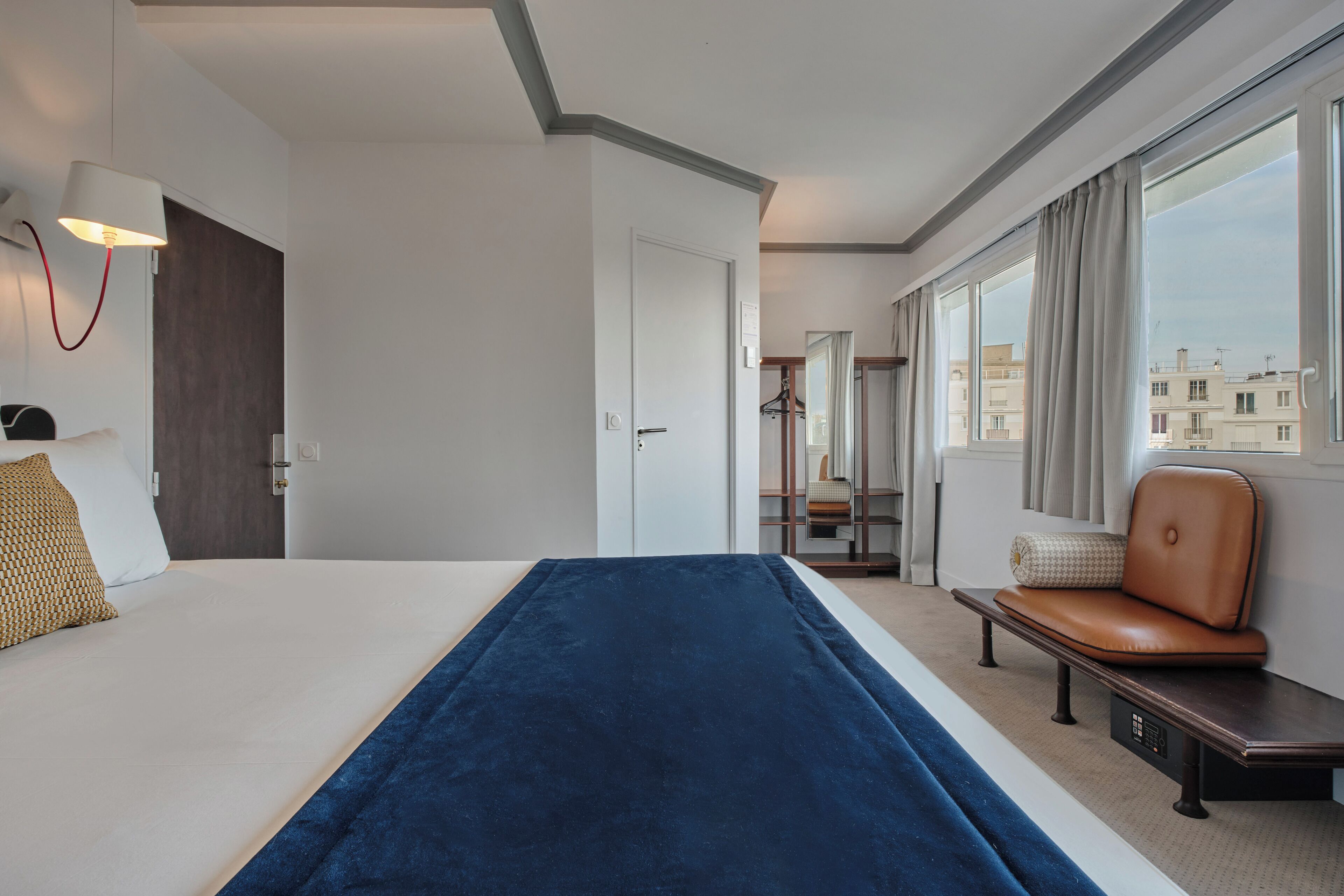 A hotel room with a bed, a bench, a desk, a lamp, a window with curtains, and a view of buildings.