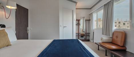 Habitación de hotel con cama, banco, escritorio, lámpara, ventana con cortinas y vistas a edificios.
