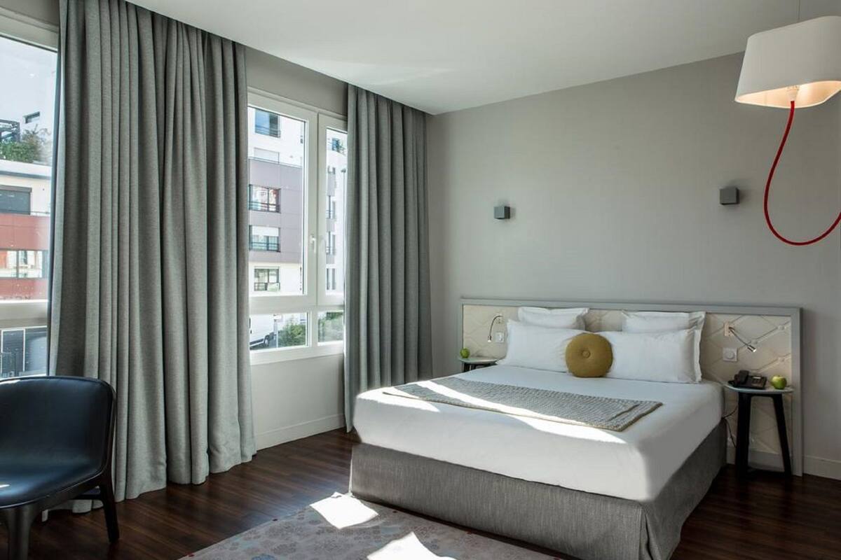 A hotel room with a large bed, a chair, a lamp, and a view of buildings outside the window.