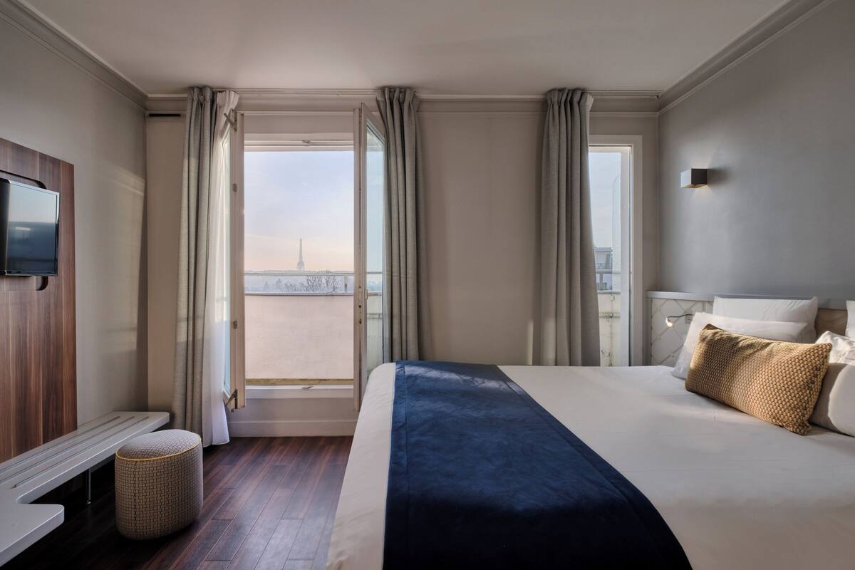 A hotel room with a bed, a TV, a bench, and a view of a cityscape.