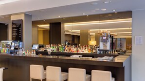 Een moderne bar met een lange toonbank, diverse drankflessen, een koffiemachine en drie barkrukken.