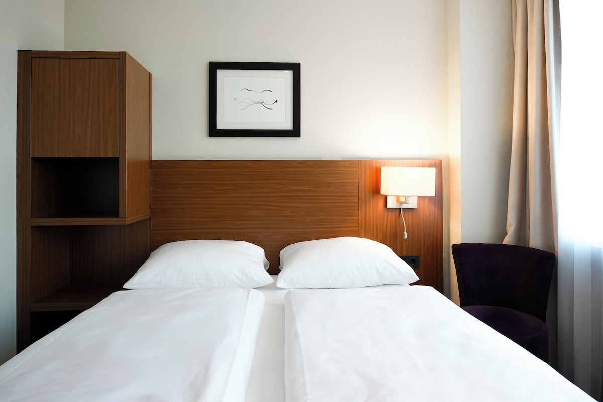 Ein Doppelbett mit weißen Leinentüchern, ein hölzerner Kopfteil, ein gerahmtes abstraktes Kunstwerk, eine Nachttischlampe und ein violetter Sessel.