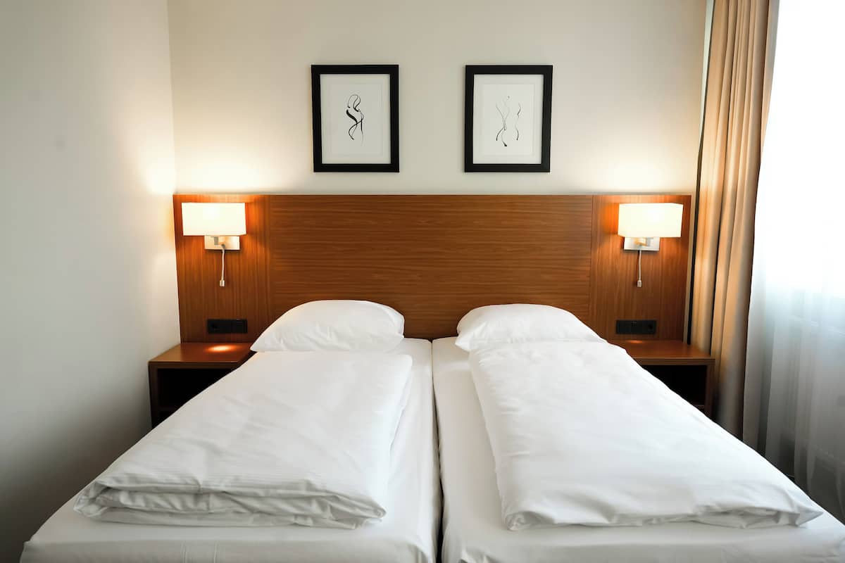 Ein Hotelzimmer mit zwei Einzelbetten, einem Holzkopfteil, zwei Nachttischlampen und zwei gerahmten Kunstwerken an der Wand.