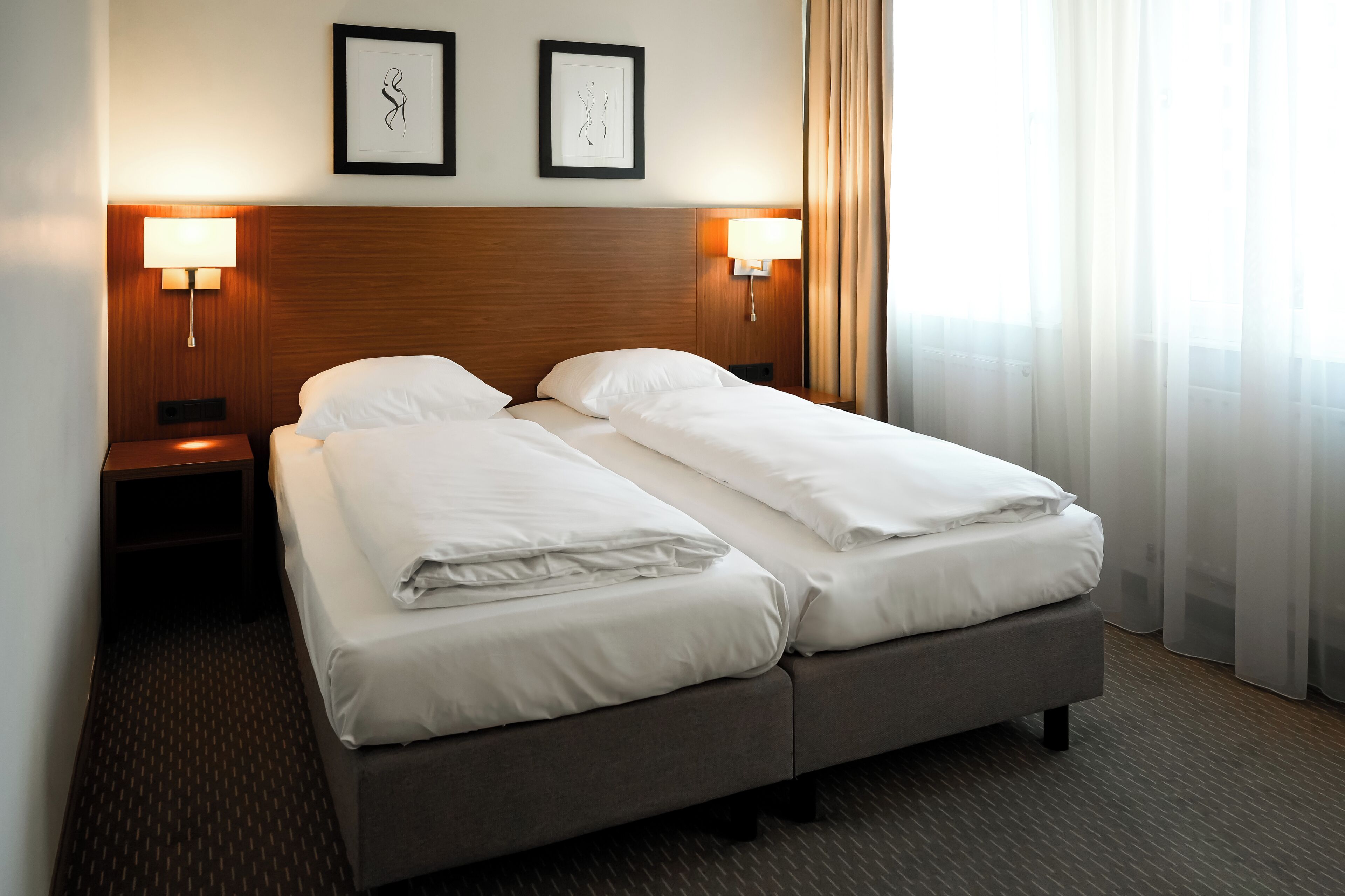A double bed with white linens, two wall-mounted lamps, and two framed pictures on the wall.