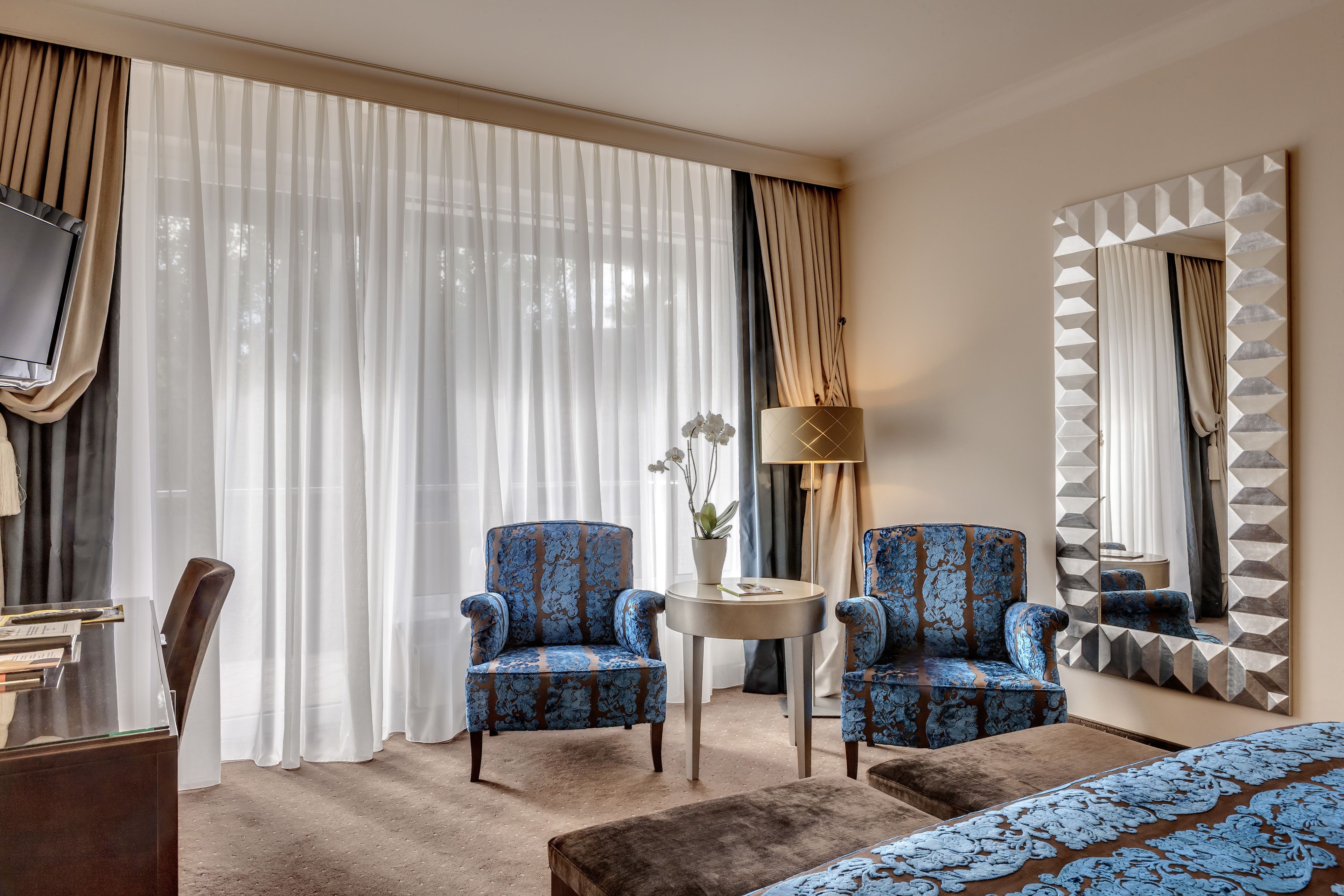 Una habitación de hotel con un ventanal grande, dos sillones, una mesita redonda con un jarrón, una lámpara, una cama y un espejo de marco geométrico.