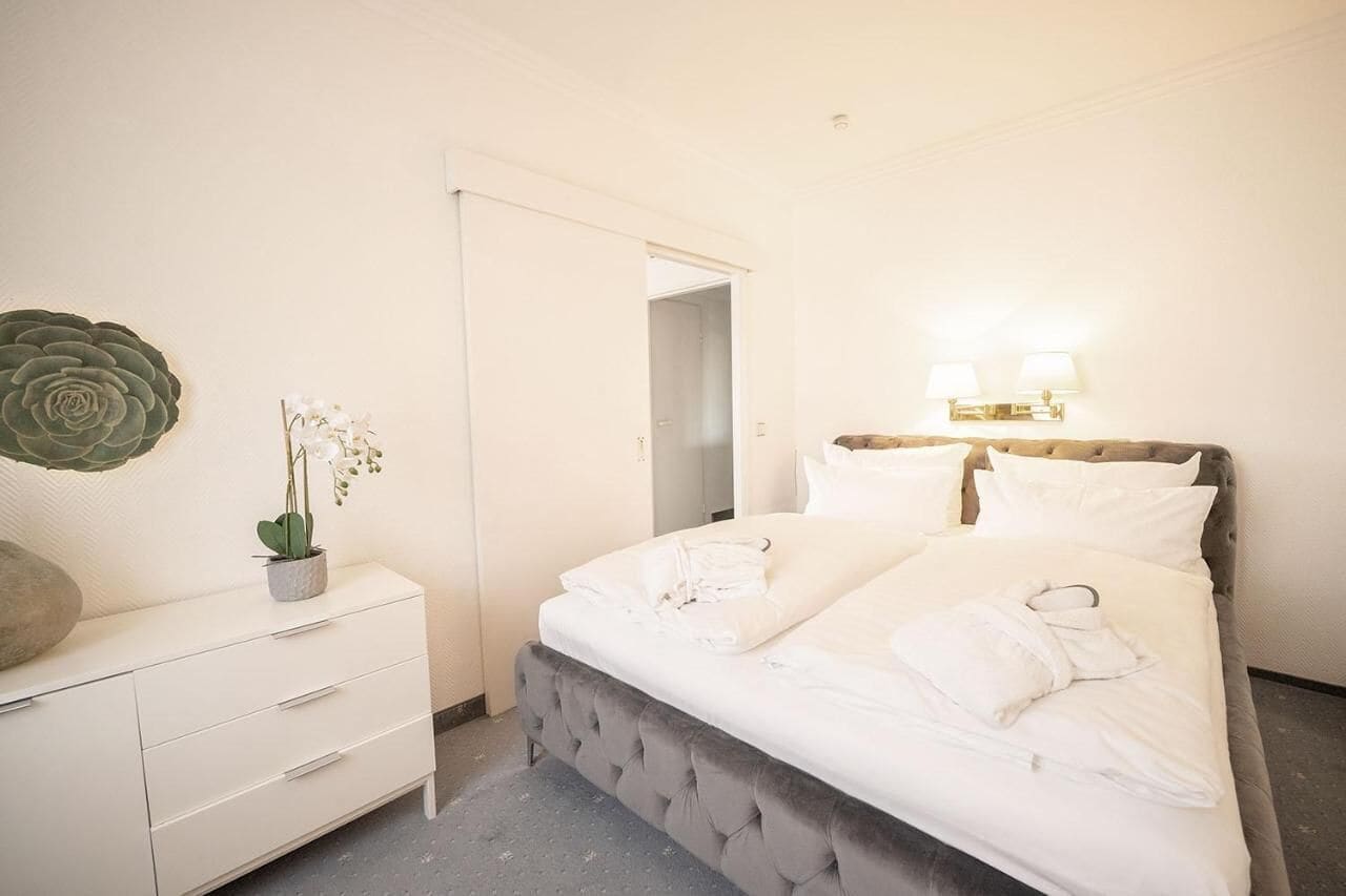 Une chambre d’hôtel avec un lit, une commode, une plante en pot et une lampe fixée au mur.