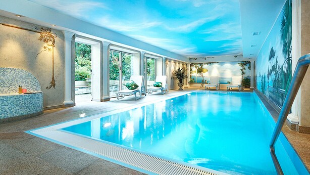 An indoor swimming pool with clear glass walls, a ceiling mural, and a seating area with a mosaic tile bench. - Bernstein Villa Kastania (Berlin)