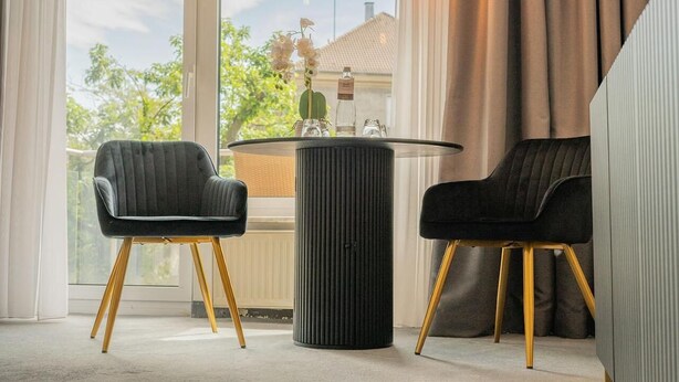 Una habitación moderna con dos sillones tapizados negros con patas de madera, una mesa redonda negra y un ventanal con cortinas.