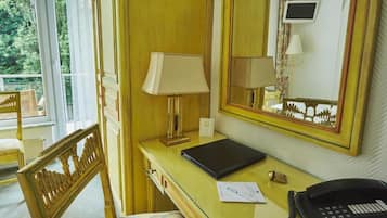 A hotel room with a wooden desk, a lamp, a telephone, a framed picture, and a mirror.