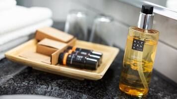 Ein Badezimmer-Waschtisch mit einer Flasche 'Rituals' Bio-Mandarine-und-Minze-Körper-Spray, einer Holzschale mit Seifenstücken und einem schwarzen Gegenstand, möglicherweise ein Rasiermesser, sowie einem Glas Wasser.