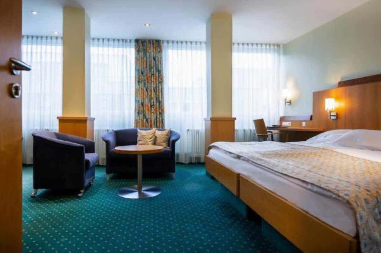 A hotel room with a bed, a round table, two armchairs, and a small desk.