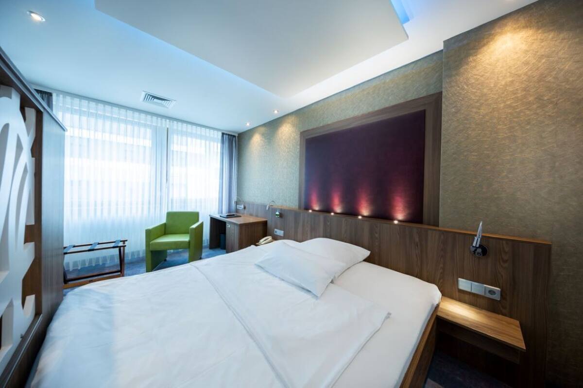 Une chambre d’hôtel avec un grand lit, un fauteuil vert, une table de chevet et un téléviseur fixé au mur.