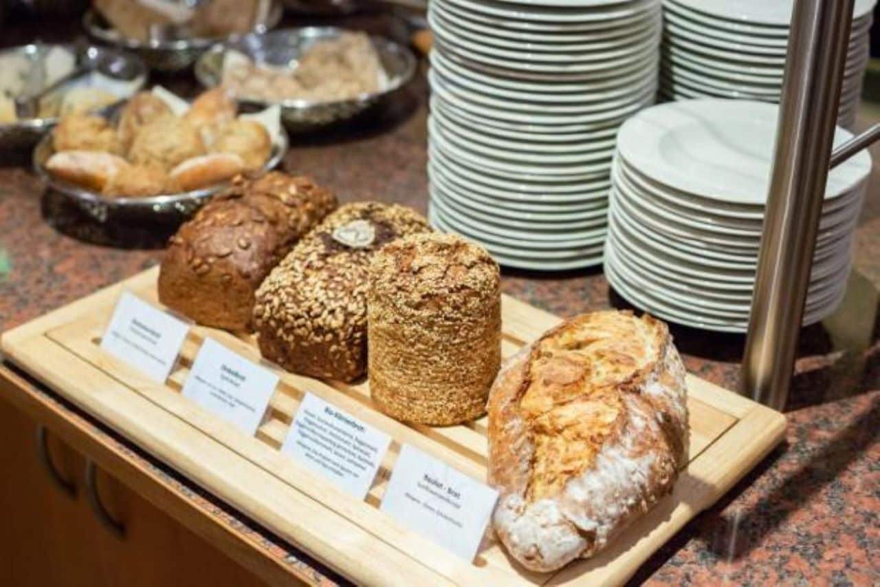 Eine Auswahl an verschiedenen Brotsorten und Gebäck auf einer Holzplatte, mit Etiketten versehen, im Hintergrund gestapelte weiße Teller.
