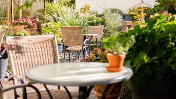 Eine Terrasse mit Korbstühlen und einem runden Tisch, umgeben von Blumentöpfen und viel Grün.