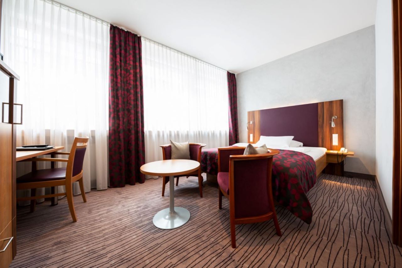 Hotelkamer met een bed, een bureau, een stoel, een ronde tafel en een groot raam met gordijnen.