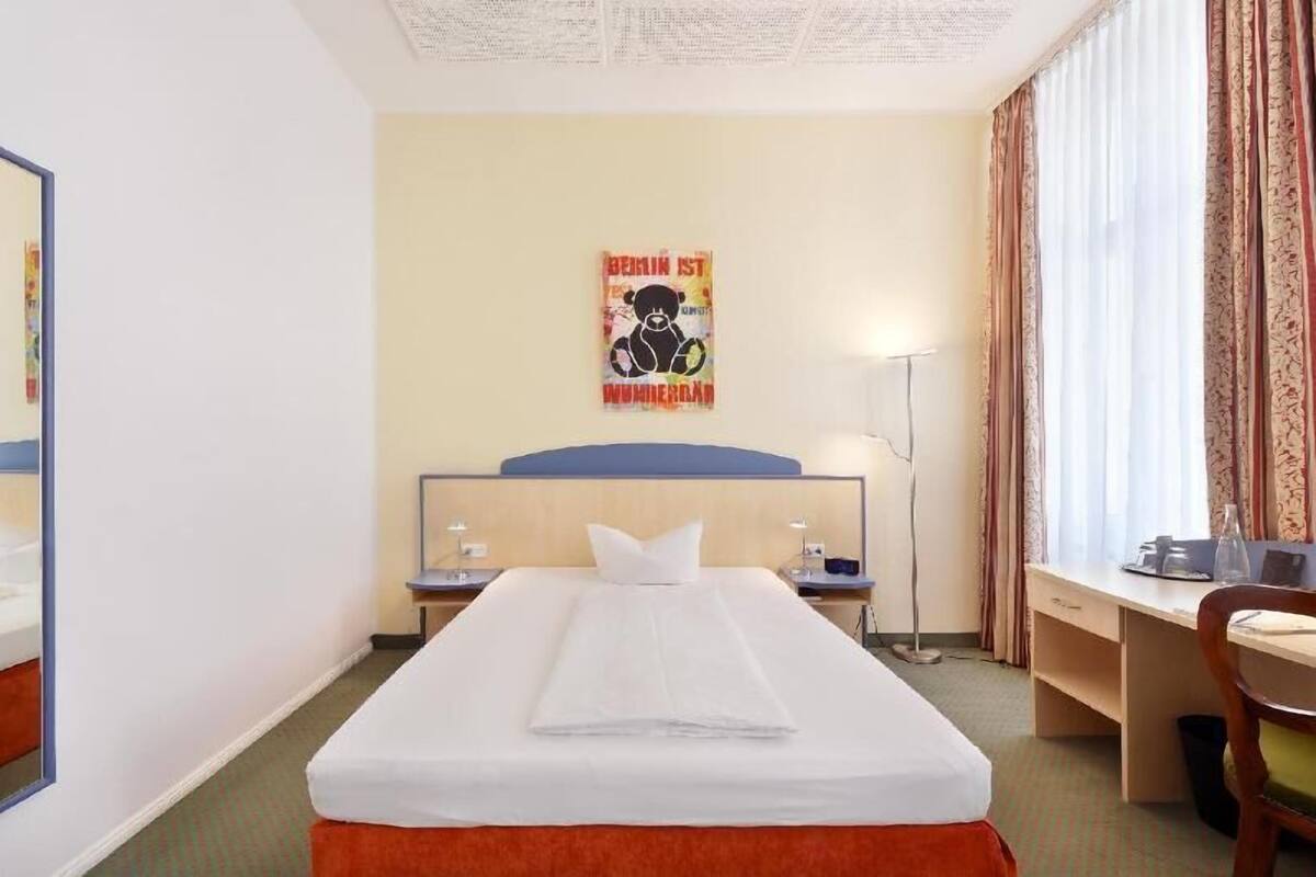 A hotel room with a bed, a desk, a chair, a lamp, and a poster on the wall.