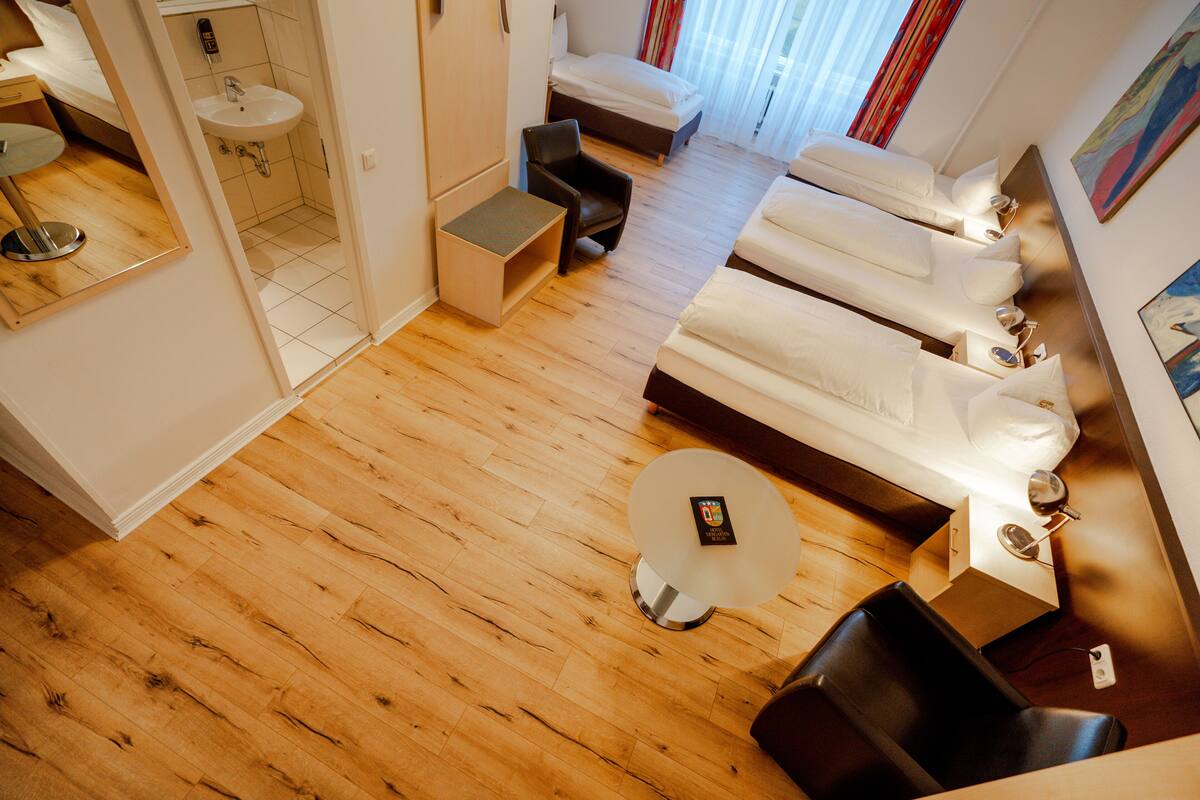 Ein modernes Hotelzimmer mit Holzboden, einem runden weißen Tisch, einer Sofagarnitur, einem kleinen weißen Waschbecken und einem Spiegel.