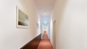 A long, narrow hallway with white walls and a red carpet. There is a framed picture on the left wall and a door at the end of the hallway.