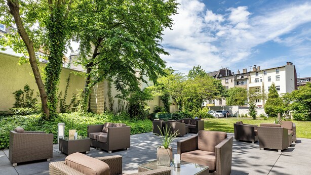 An outdoor patio area with wicker furniture and a glass table, surrounded by greenery and a building in the background. - Garner Hotel Bochum by IHG (Bochum)