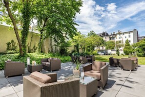 An outdoor patio area with wicker furniture and a glass table, surrounded by greenery and a building in the background. - Garner Hotel Bochum by IHG (Bochum)