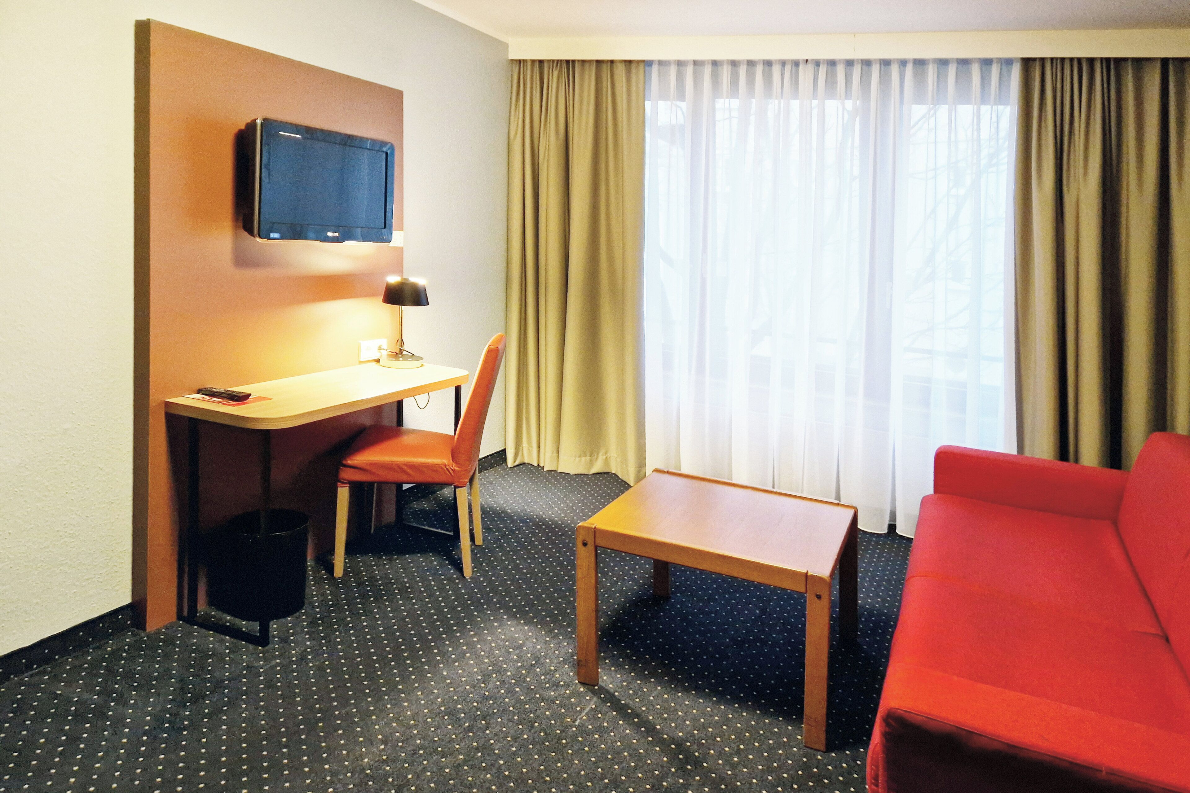 A hotel room with a desk, chair, TV, red sofa, and a window with curtains.