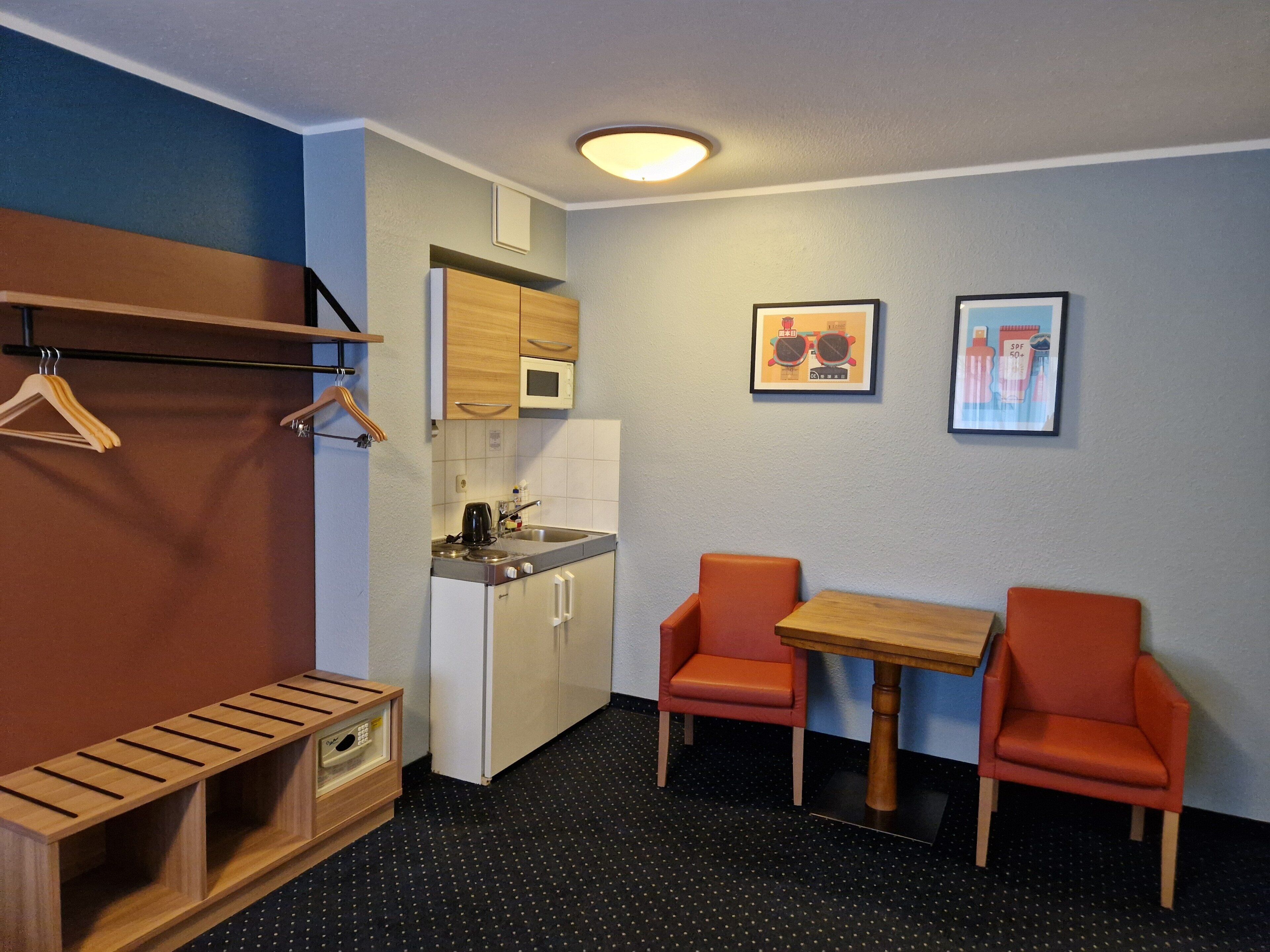 Une cuisine compacte équipée d’un four à micro-ondes, d’un évier et de meubles en bois. On y trouve deux chaises orange et une petite table.