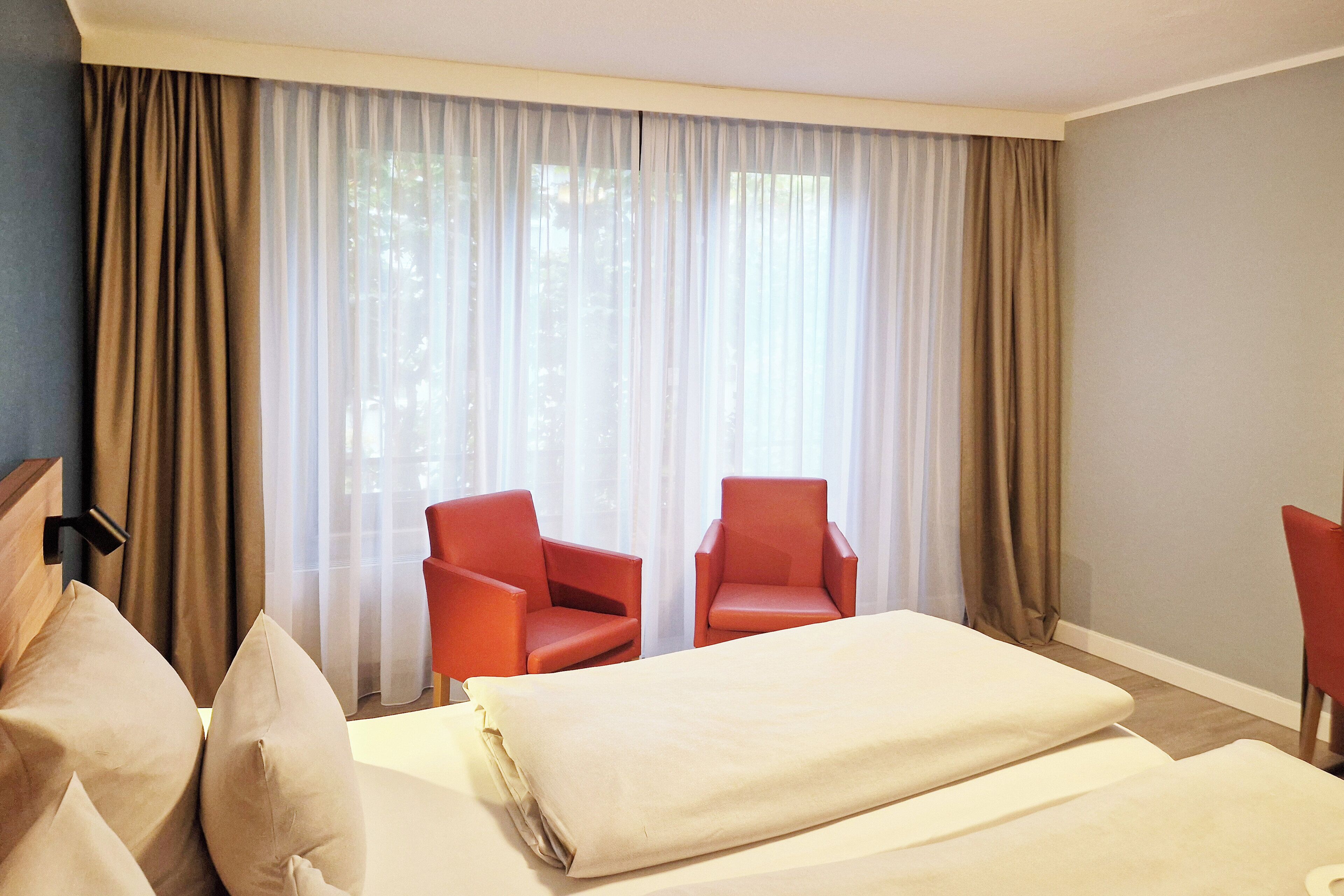 Ein Hotelzimmer mit zwei roten Sesseln, einem Bett mit weißer Bettwäsche und durchsichtigen Vorhängen an den Fenstern.