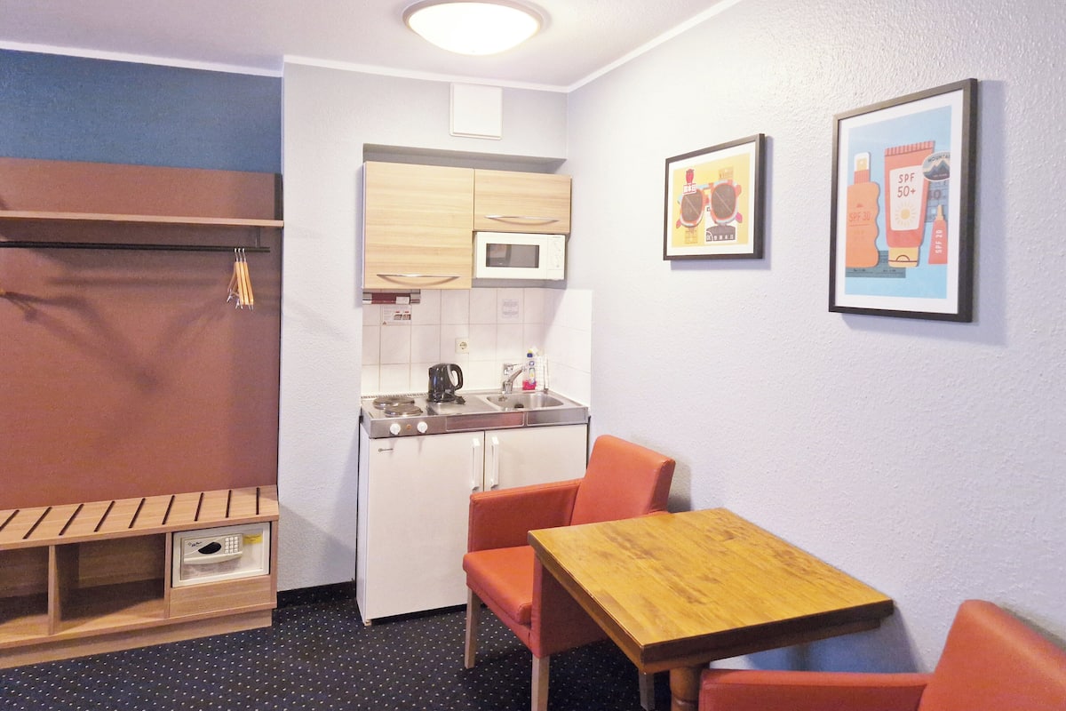 Een compacte keuken met een magnetron, spoelbak en houten kastjes. Er is een kleine eettafel met twee stoelen en een bankje.