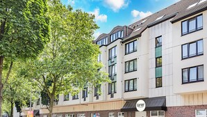 A multi-story building with a parking sign, a storefront with a sign, and a tree in front. - Garner Hotel Bochum by IHG (Bochum)