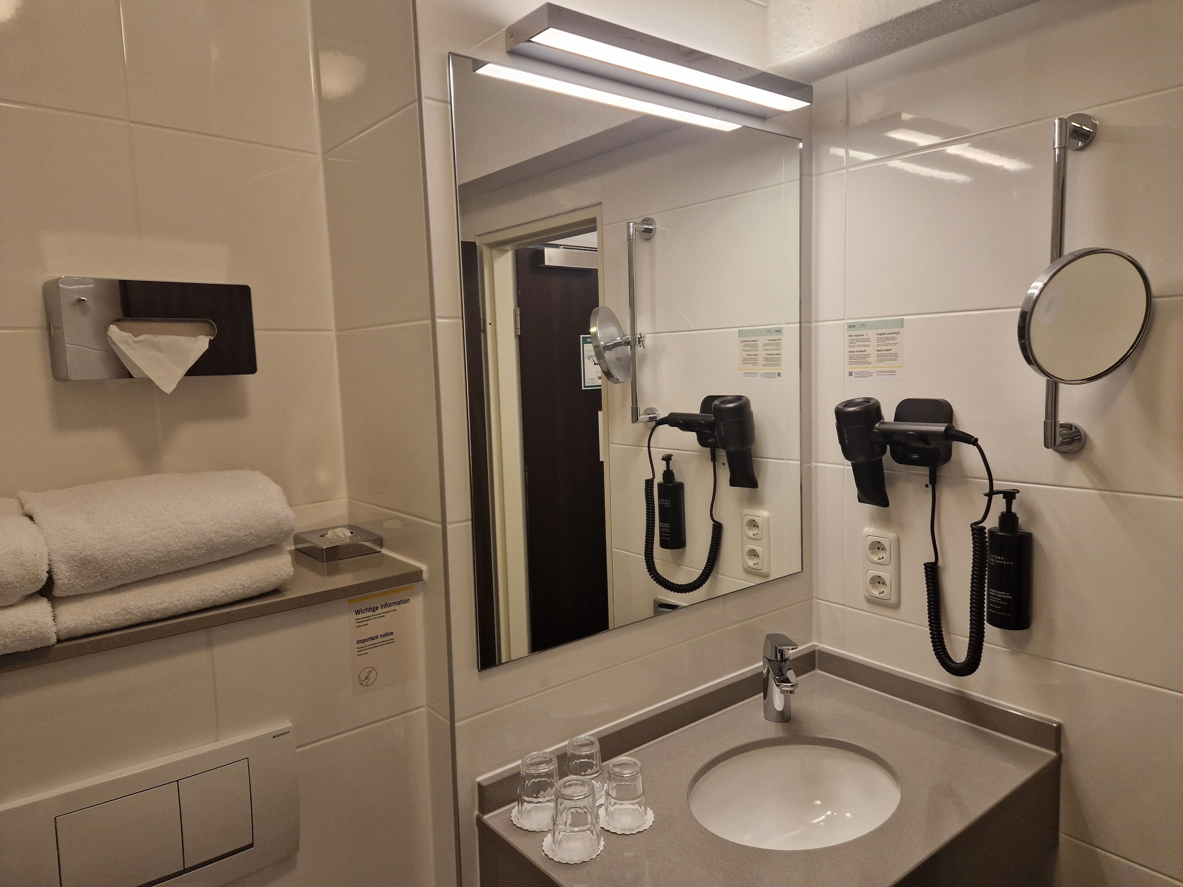 A hotel bathroom with a sink, mirror, towel dispenser, and hair dryers.