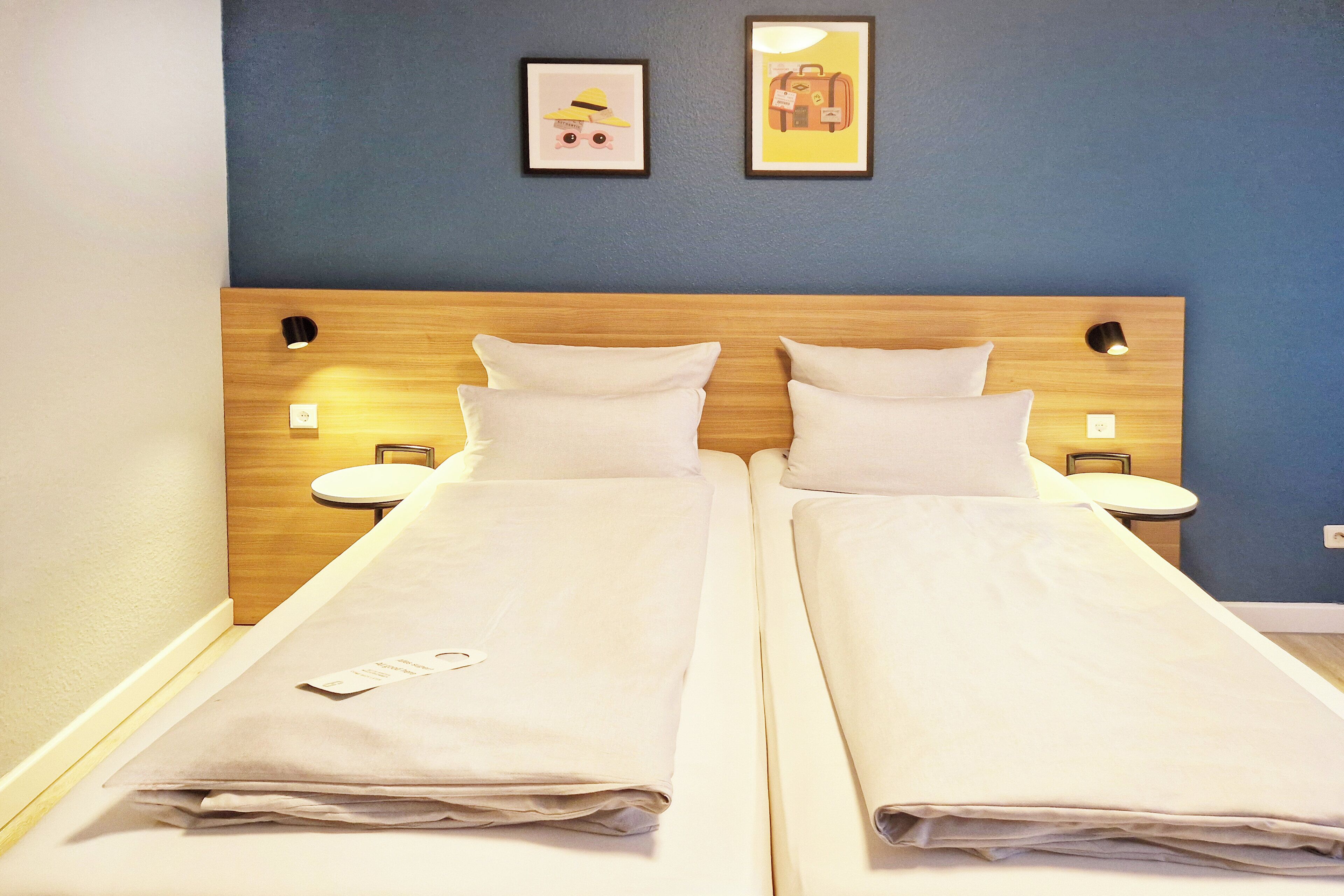 Una cama doble con ropa de cama blanca, dos mesitas de noche con lámparas y dos cuadros en la pared.