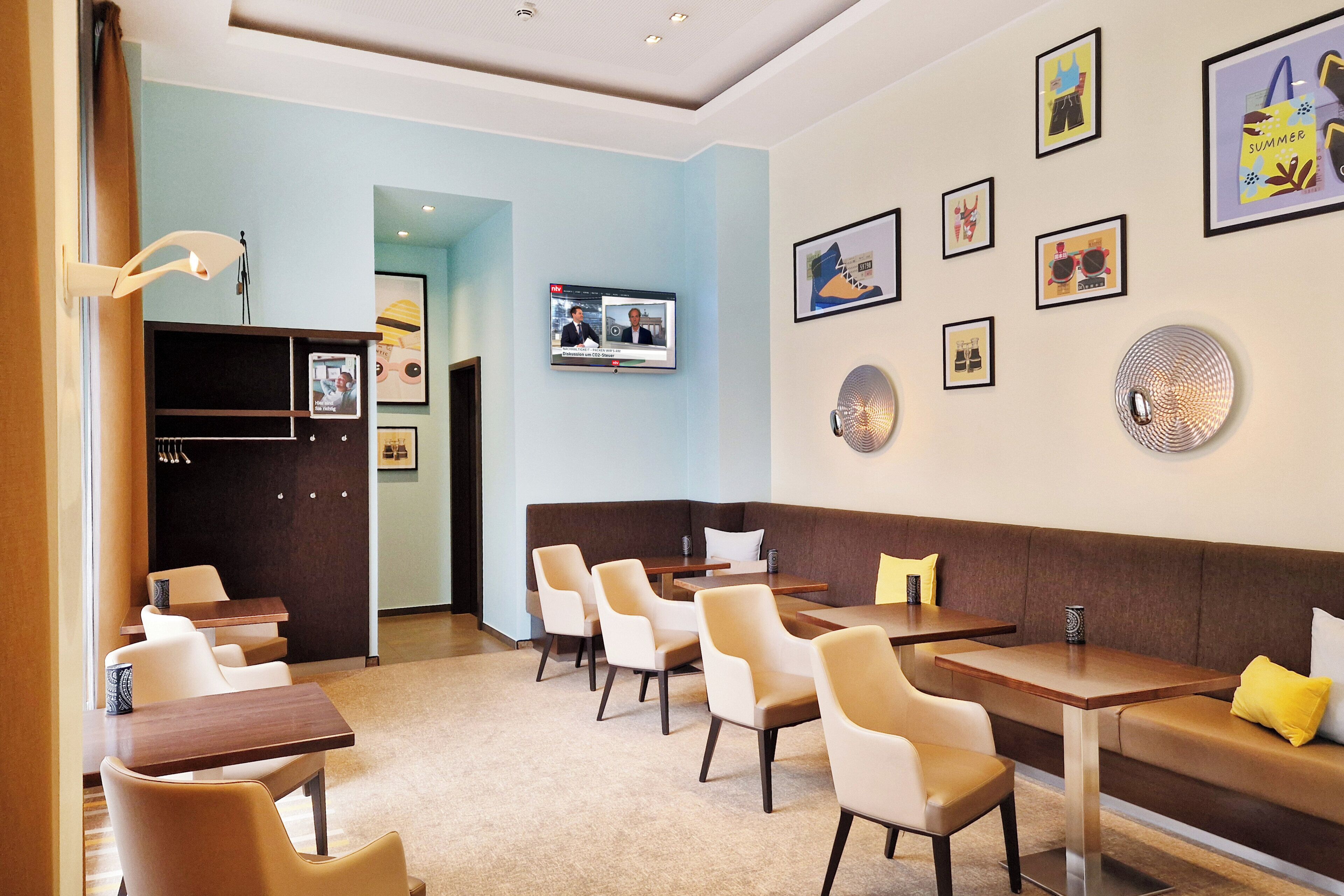 Een moderne hotellobby met een zithoek, een televisie waarop een nieuwsuitzending wordt vertoond, en diverse ingelijste foto's aan de wanden.