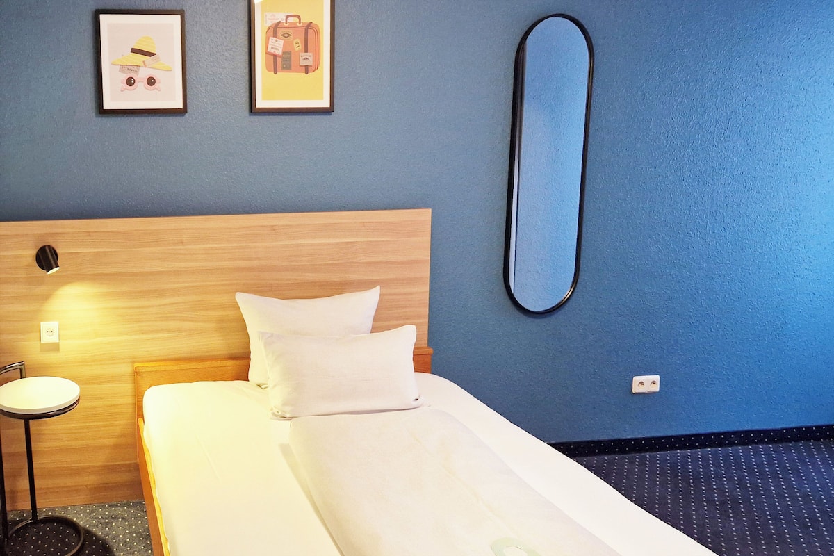 Een hotelkamer met een houten hoofdeinde, een enkel bed met wit beddengoed, een ronde spiegel aan de muur en twee ingelijste foto's boven het bed.