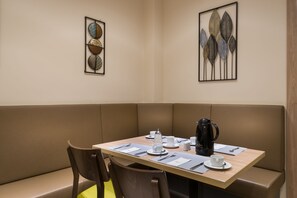 Een zithoek met eethoek, met een houten tafel gedekt voor twee personen, een bruine gestoffeerde bank en twee lijstwerken aan de muur.