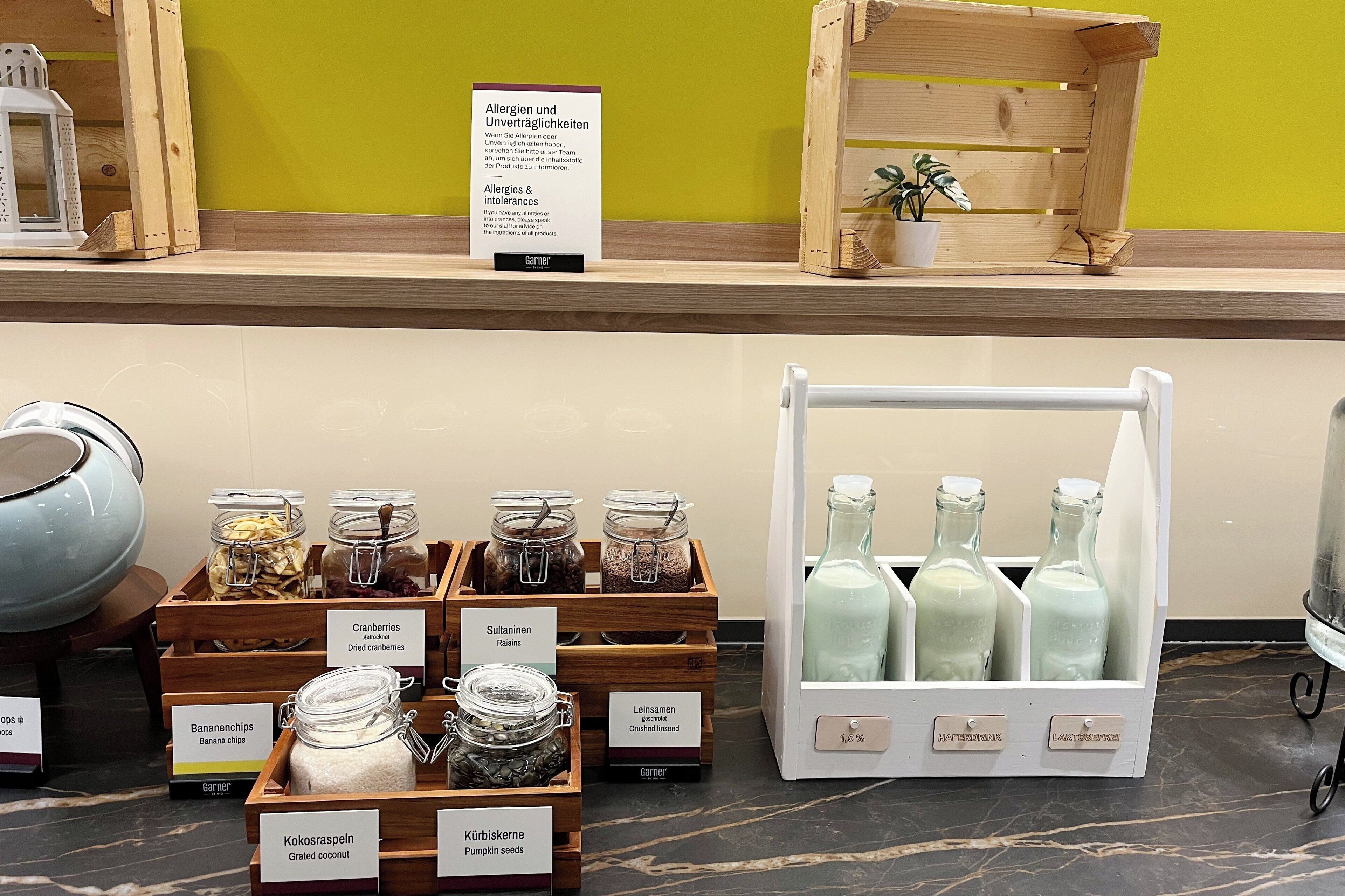 A display of various food items and beverages in wooden crates and glass jars, with labels indicating contents like cranberries, sultanas, and coconut.