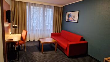 A hotel room with a red sofa, a wooden coffee table, a desk with a lamp, and a TV mounted on the wall.