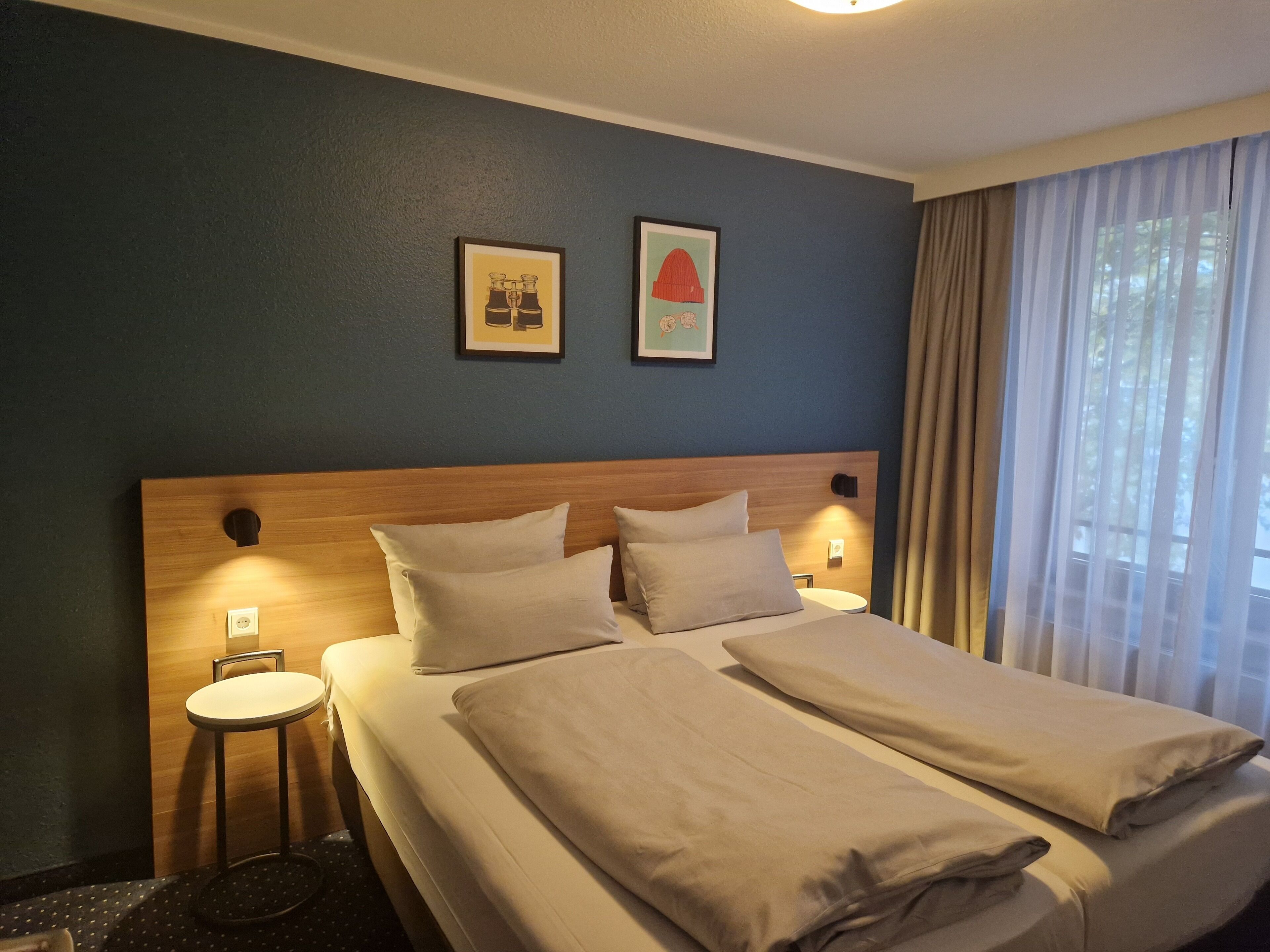 A hotel room with a double bed, two pillows, a small bedside table, and a wall-mounted lamp.