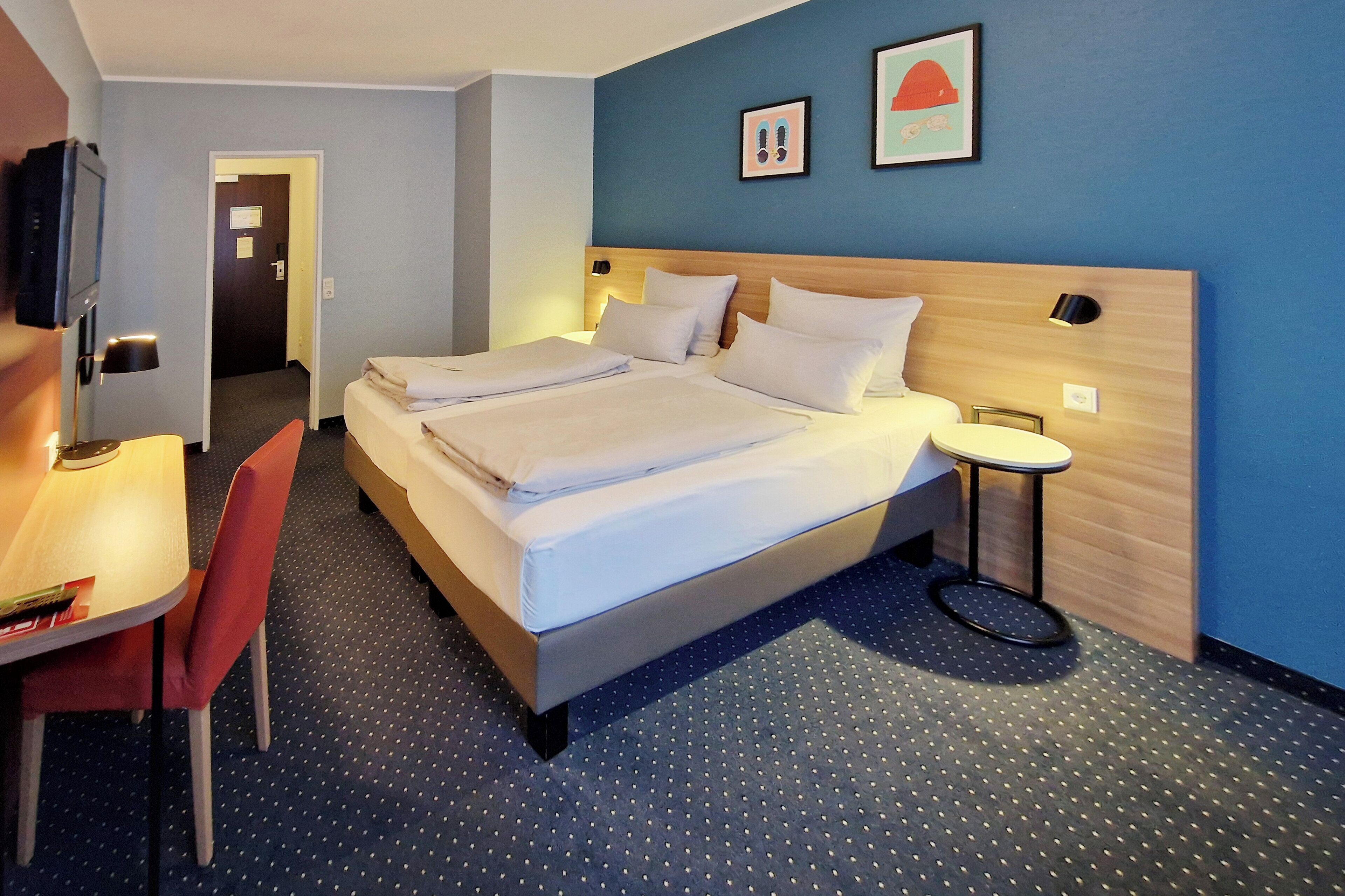 A hotel room with a bed, a desk, and a chair. There is a door leading to another room.