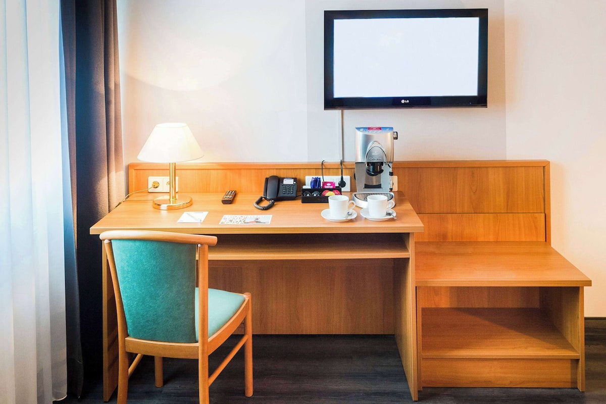A hotel room with a wooden desk, a teal chair, a coffee maker, a phone, and a TV on the wall.