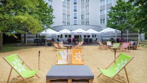 An outdoor seating area with beach chairs and tables, umbrellas, and a building in the background. - Hotel Bochum Wattenscheid Affiliated by Meliá (Bochum)