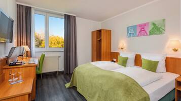 A hotel room with a bed, a desk with a computer, a chair, a window with curtains, and a painting on the wall.