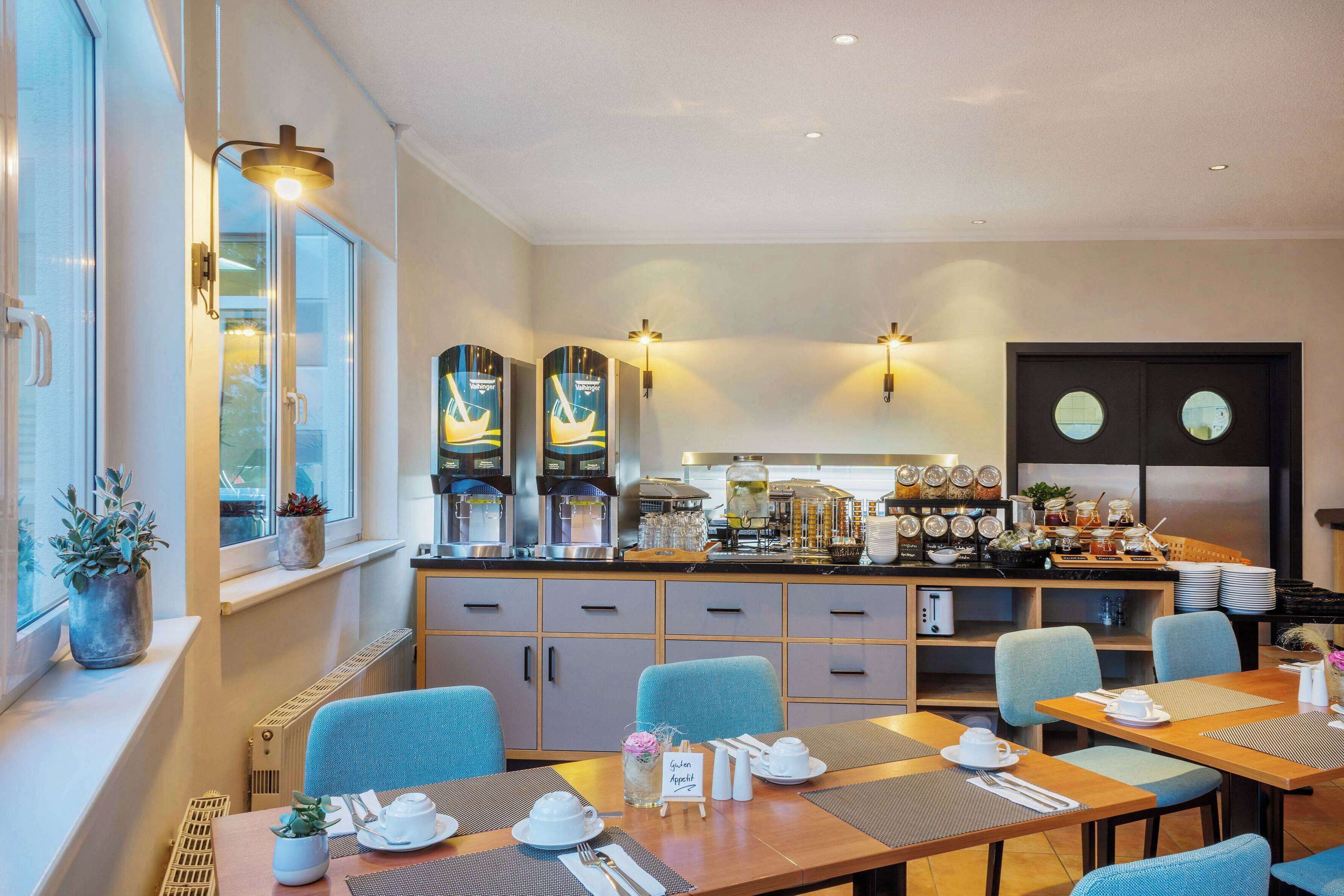 A modern hotel dining area with a long counter, multiple seating options, and a kitchen in the background.