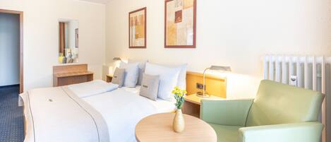 Una habitación de hotel con una cama, una mesita de noche con lámpara, una silla, una pequeña mesa con un florero y una obra de arte en la pared.