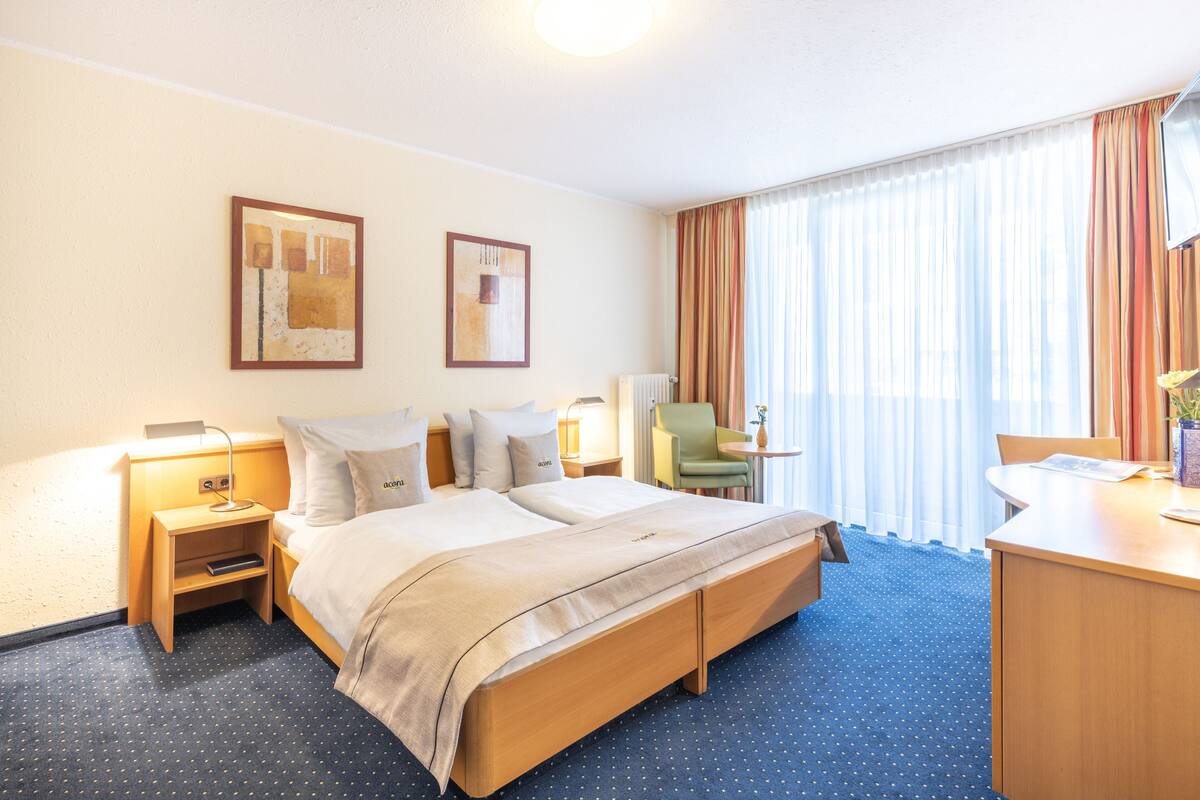 A hotel room with a large bed, two bedside tables with lamps, a desk with a chair, and a window with curtains.