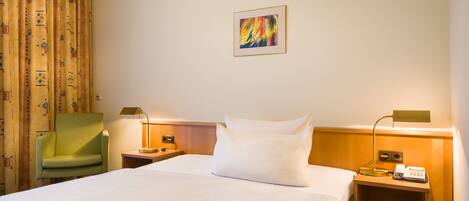 A hotel room with a bed, a green armchair, a nightstand with a lamp, a phone, and a painting on the wall.