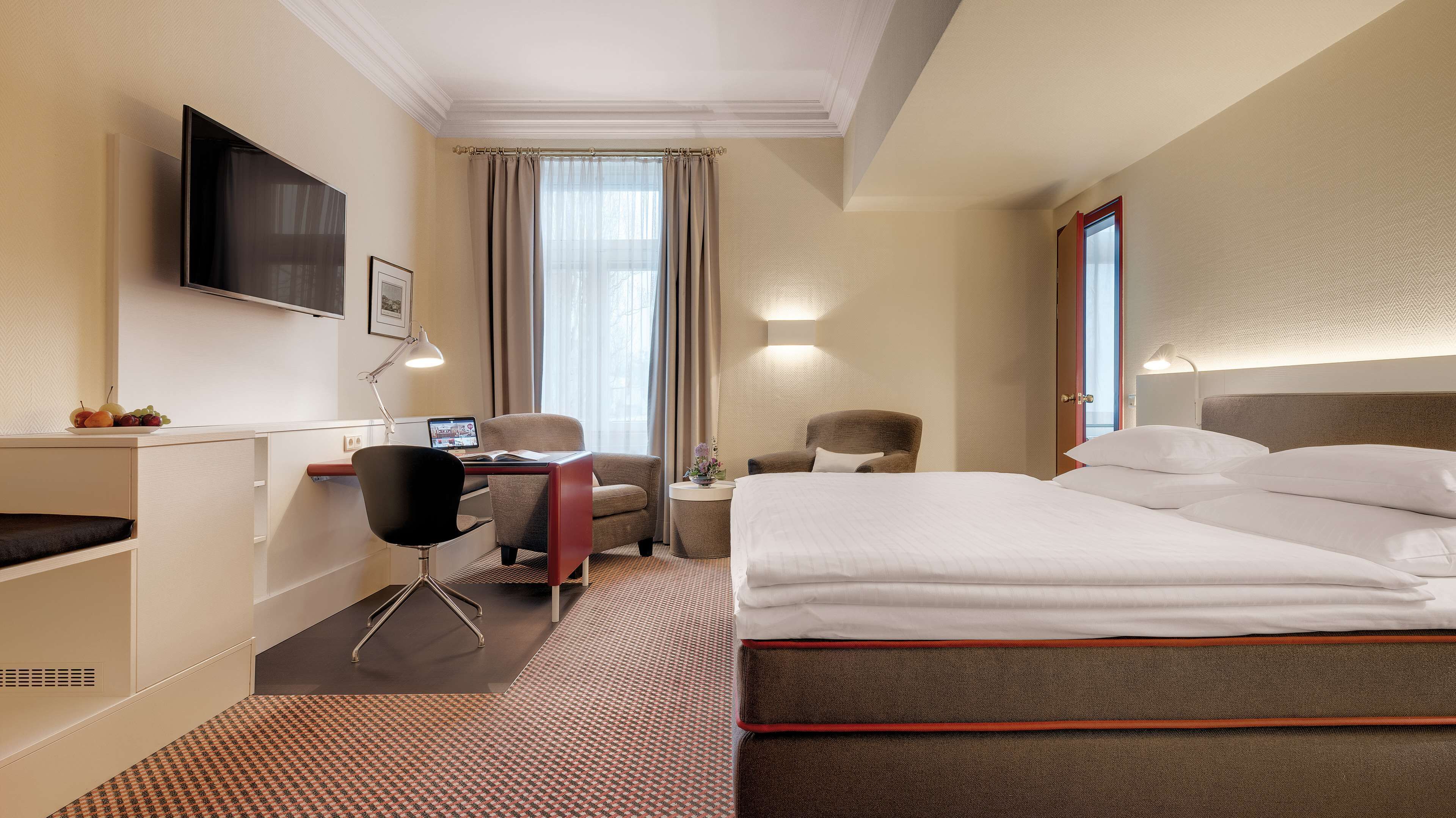 A hotel room with a bed, desk, chair, TV, and a window with curtains.